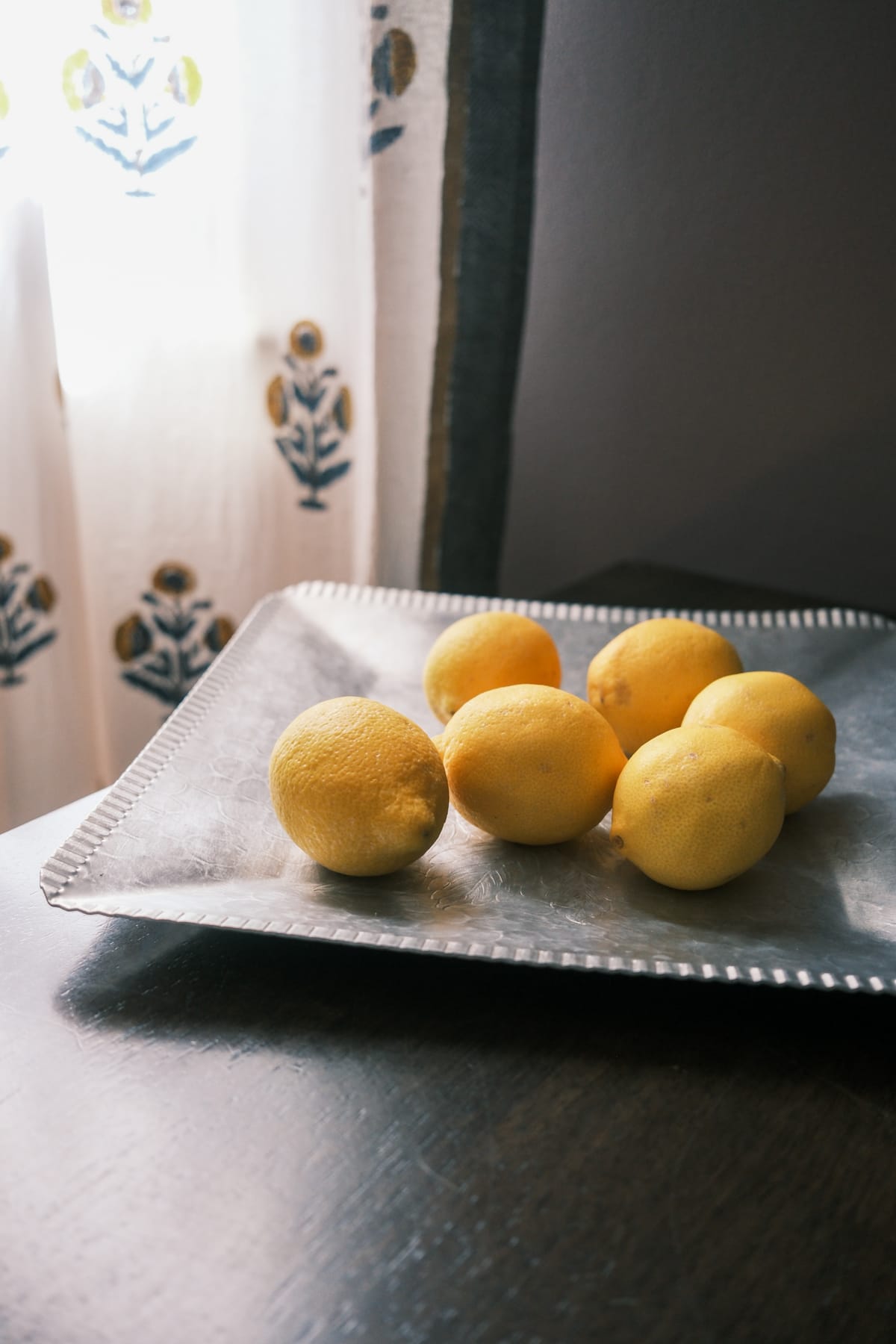 Six lemons on a hammered pewter tray beside a window, with a blue and ochre block-print curtain filtering the light