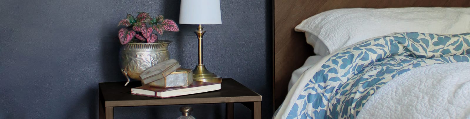 A styled bedroom with deep navy walls, a brass lamp and books on a bedside table, and a blue floral quilt on the bed