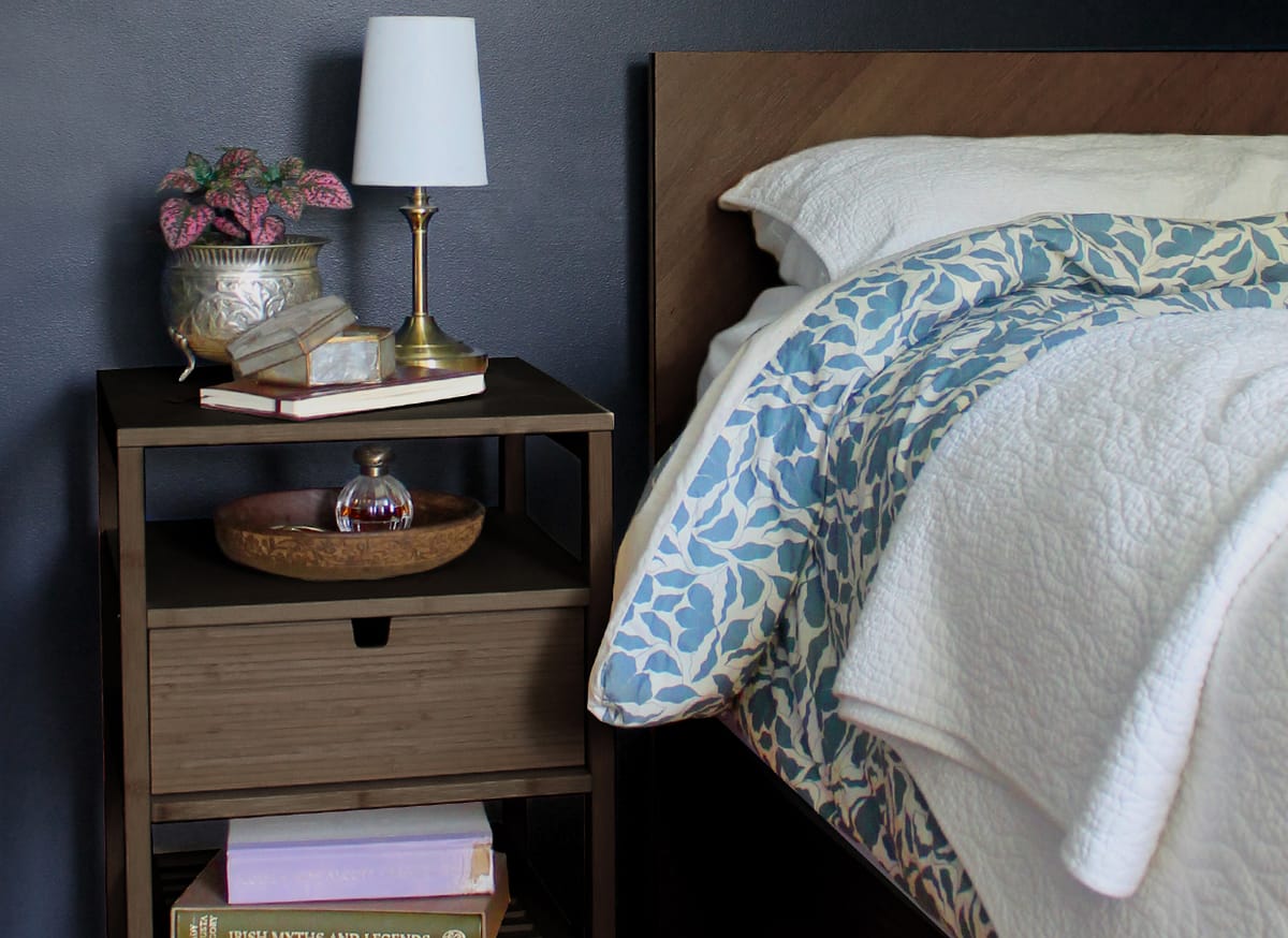 A bedroom nightstand styled with a brass lamp, books and a plant, beside a blue floral quilt